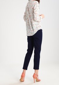 Blouse dengan motif bunga dan detail kancing, celana navy, dan sandal hak tinggi berwarna koral. Kain halus, potongan pas, panjang hingga pergelangan kaki.