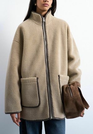 Donna che indossa una giacca in pile beige con grandi tasche frontali e tiene una borsa marrone in camoscio con chiusura a zip per il manico.