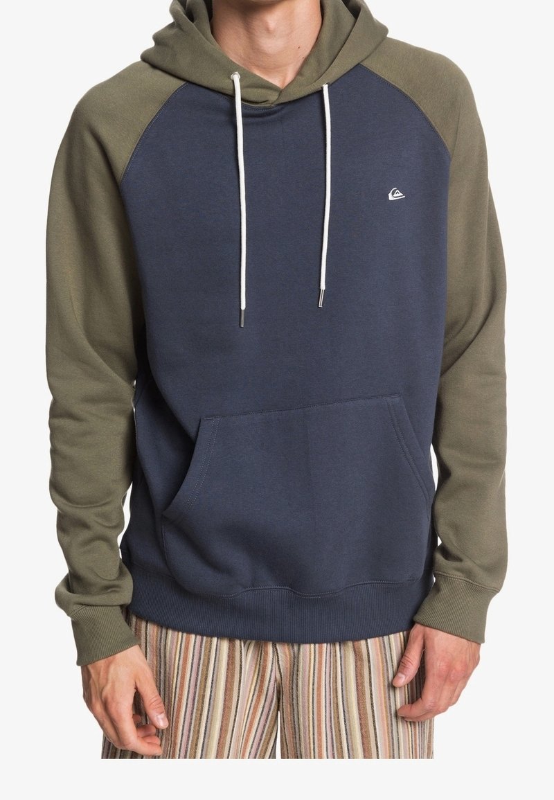 Homme portant un sweat à capuche bleu marine avec des manches vert olive et des cordons blancs, associé à un pantalon rayé multicolore.