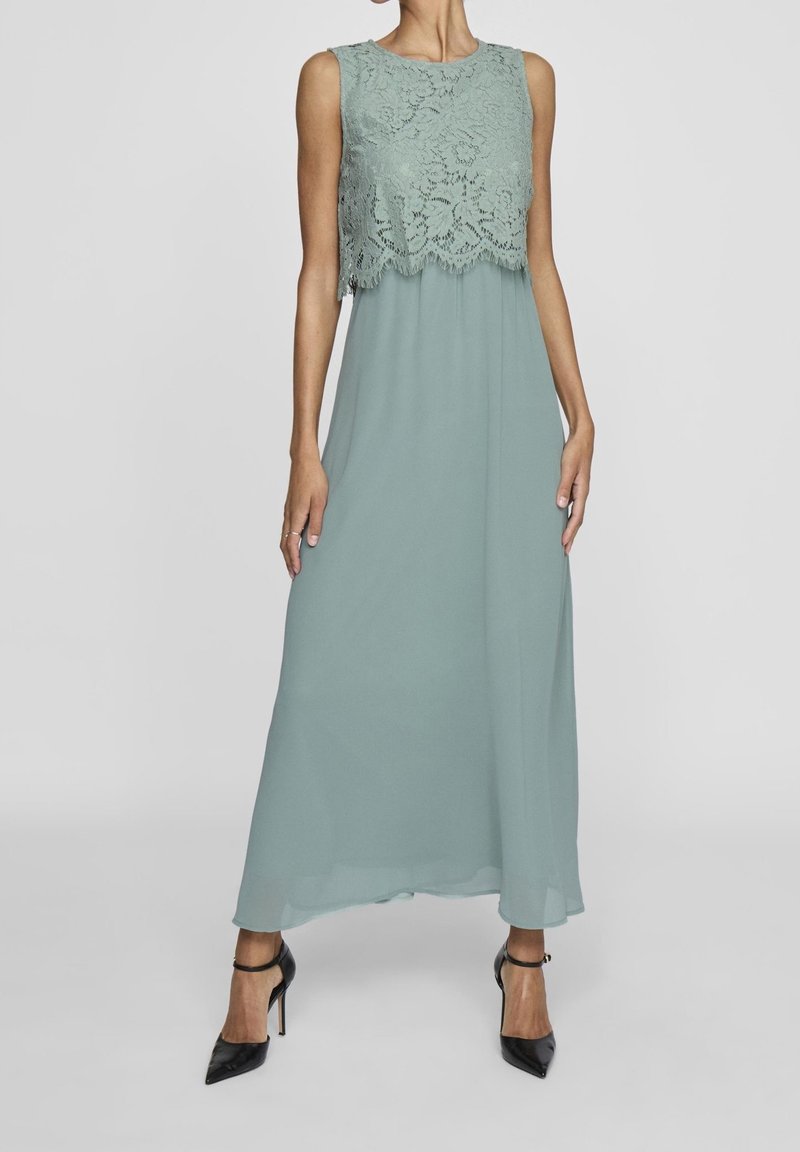 Femme portant une robe maxi vert sauge sans manches avec un corsage en dentelle superposée et des talons hauts noirs à bride cheville, debout devant un fond uni.
