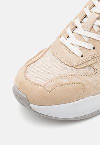 Beige mockasinsneaker med texturerad tyg framdel, vita snören och vit gummisula mot en enkel vit bakgrund.