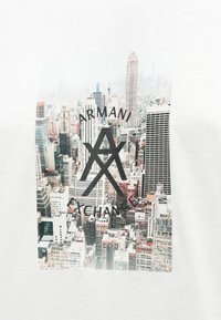 Balta T-kreksa ar fotogrāfisku pilsētas siluetu un melnu "Armani Exchange" logotipu ar ģeometriskiem akcentiem.