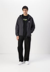 Graue, leichte Jacke mit Kapuze, Reißverschluss vorne und seitlichen Taschen. Schwarzes T-Shirt mit grünem Grafiktext, kombiniert mit schwarzen Hosen und weißen Sneakern.