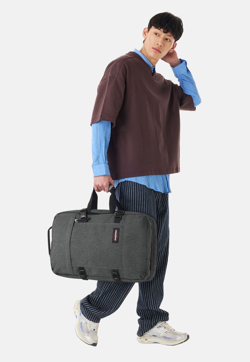 Jeune homme portant un grand sac de voyage gris, vêtu d’une chemise marron à manches courtes sur une chemise bleue à manches longues et un pantalon rayé.