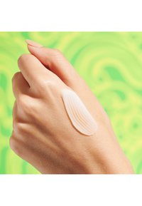 Moisturizing cream spread on light brown hand, smooth texture, off-white color, against a vibrant green background.