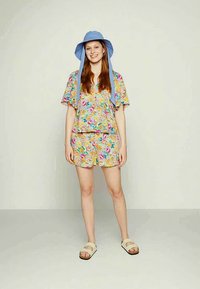 Conjunto floral de duas peças com uma blusa de mangas curtas e calções em cores vibrantes. Acompanhado por um chapéu de aba larga azul claro e sandálias.