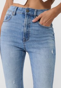 Blaue Skinny-Jeans aus Denim mit hoher Taille, mit einem fransigen Detail über dem Knie, fünf Taschen und einem Metallknopfverschluss.