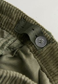 Olivgrüner Corduroy-Taillengürtel mit texturiertem Muster, ausgestattet mit einem flachen Metallknopf und einem gewebten Etikett. Das Innenfutter ist aus glattem Stoff.