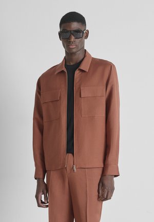 Man met een bruin jasje en broek, zwarte shirt en grote zwarte zonnebril, staande tegen een effen lichte achtergrond.