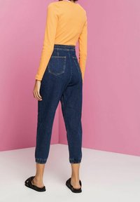 Denim jogger-housut, joissa on joustava vyötärö, kapenevat hihnat ja kaksi takataskua; päällä pitkähihaisen oranssin paidan kanssa ja mustat sandaalit.