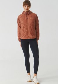 Veste matelassée couleur rouille avec capuche, fermeture éclair sur le devant et poches à rabat, associée à des leggings navy et des baskets blanches.