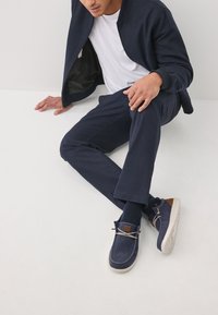 Navyblaue Sneaker aus Stoff mit weißer, strukturierter Sohle, braunen Lederakzenten und beigen Schnürsenkeln; getragen zu navyblauen Hosen und dunklen Socken.