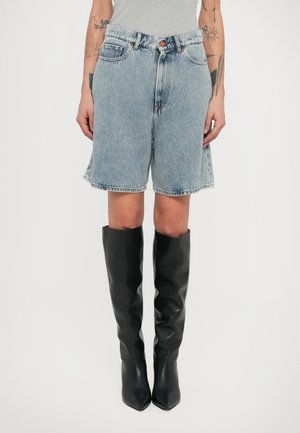 Osoba ubrana w jasnoniebieskie szorty jeansowe i czarne kozaki sięgające do kolan, z widocznymi tatuażami na przedramionach, na jednolitym białym tle.