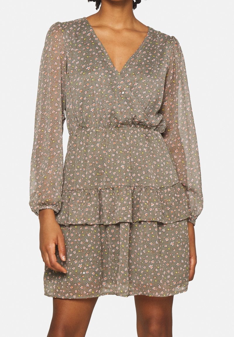 Femme portant une robe longue à manches longues verte à motifs floraux, avec un décolleté en V, une taille cintrée et une jupe à volants à étages.