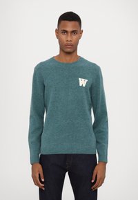Merino wool sweater in teal with a round neck, long sleeves, and a white "W" emblem on the left chest. Ribbed hem and cuffs.