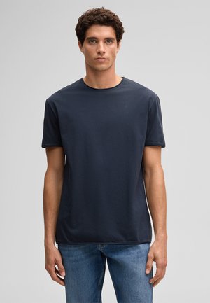 Jeune homme aux cheveux bouclés et foncés, portant un t-shirt à manches courtes bleu foncé uni et un jean bleu, debout devant un fond gris.