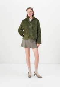 Vrouw gekleed in een olijfgroen nep bont zipjack, geruite minirok, beige sokken en taupe puntige hakken, staand tegen een witte achtergrond.