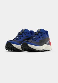 Um par de sapatilhas de trail running azuis e pretas com entressolas brancas, atacadores pretos e detalhes vermelhos nos calcanhares, num fundo branco.