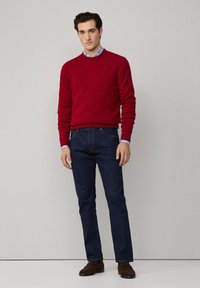 Un pull en maille torsadée rouge, une chemise bleue à col en dessous, un jean en denim foncé et des chaussures en suède marron. Le mannequin se tient devant un fond uni.