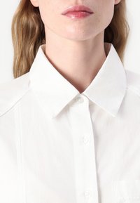 White button-up shirt with a pointed collar, made of smooth fabric, featuring a pocket and tonal stitching along the placket.