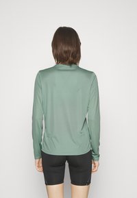 Langarm-Sportoberteil in gedämpftem Grün, aus glattem, leichtem Stoff. Mit einem Rundhalsausschnitt und tailliertem Design. Kombination mit schwarzen Shorts.