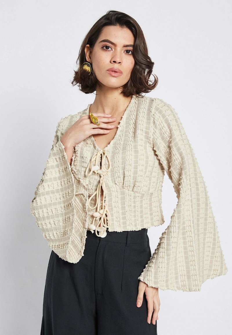 Beige top cropped dengan kain bertekstur, leher V, dan lengan mengembang. Memiliki bagian depan dengan tali yang dapat diikat dan hiasan manik-manik.