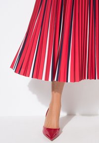 Jupe plissée rouge avec des rayures blanches et marine au bas, associée à des escarpins rouges brillants à bout pointu. Accent sur la texture et le contraste de couleurs vives.