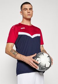 Rood- en marineblauw teamshirt met korte mouwen, met een wit zigzagpatroon en logo. Speler houdt een zwart-witte voetbal met patroon vast.