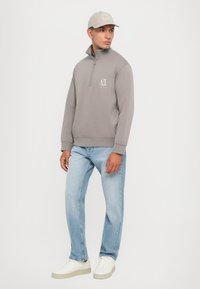 Sudadera gris con cremallera y un pequeño logotipo, combinada con vaqueros de mezclilla azul claro y zapatillas blancas. Los accesorios incluyen una gorra gris claro.