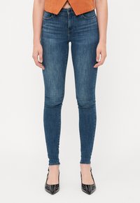A vékony farmerek sötétkék denim anyagból készültek, közepes derékmagassággal, elülső zsebekkel és sima felülettel rendelkeznek. Fekete, hegyes sarkú cipővel viselték.