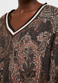 Pull en laine de couleur marron avec un motif, présentant un décolleté en V et des détails en contraste noir et crème au niveau du col côtelé. Tissu doux et texturé.