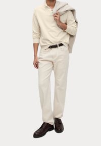 Mann in cremefarbenem langärmeligen Polo, passenden Hosen, schwarzem Gürtel, dunkelbraunen Schnürschuhen und hält einen beigen Pullover über der Schulter.