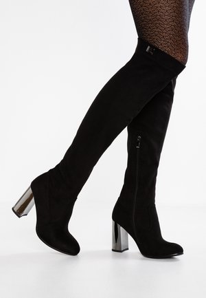 Czarne zamszowe kozaki za kolano z srebrnymi słupkowymi obcasami, noszone z wzorzystymi rajstopami, pokazujące boczny suwak oraz ułożenie stopy pod kątem.