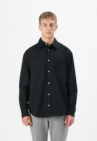 Jonge man staat, draagt een zwart overhemd met knopen en een borstzakje en lichtgrijze broek tegen een simpele witte achtergrond.
