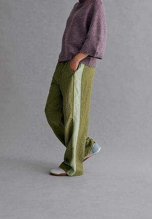 Pantalon en lin vert avec une bande latérale vert clair, associé à un pull tricoté lavande. La personne a les mains dans les poches, debout devant un fond gris.