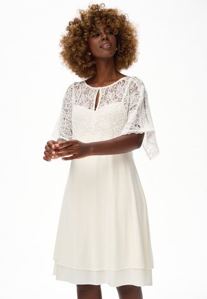 Kobieta o kręconych włosach, ubrana w białą sukienkę do kolan z kwiatowym koronkowym gorsetem i krótkimi falbankowymi rękawami, stojąca na jednolitym tle.