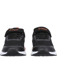 Baskets noires de sport avec une tige en maille respirante, dotées d'une sangle Velcro, d'une surface texturée et d'une semelle en caoutchouc blanche avec une bande de roulement noire.