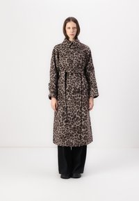 Pledd av leopardmønster i trenchcoat laget av mykt stoff, med belted liv, stor krage og dobbelte knapper. Svarte bukser er brukt under.
