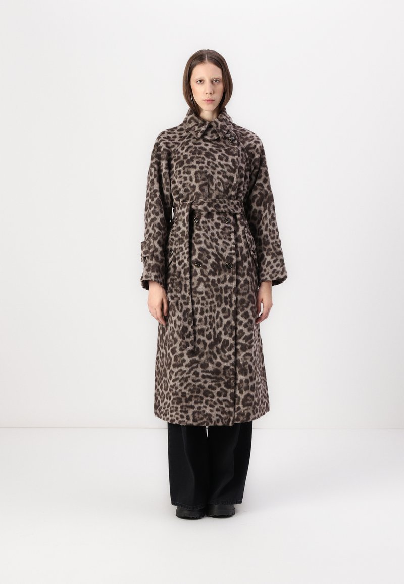 Pledd av leopardmønster i trenchcoat laget av mykt stoff, med belted liv, stor krage og dobbelte knapper. Svarte bukser er brukt under.