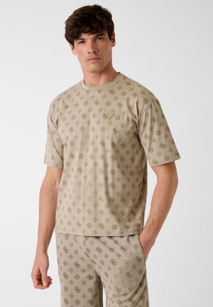 Uomo che indossa una camicia beige a maniche corte e pantaloni abbinati con un motivo geometrico scuro e logo ripetuto, in piedi davanti a uno sfondo semplice.