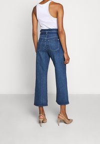 Hoge taille, wijde blauwdenim jeans met twee achterzakken en een gladde textuur, gecombineerd met een witte tanktop en neutrale hakken.