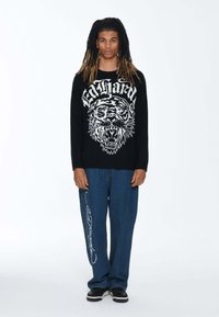 Czarny sweter z białą grafiką tygrysa i napisem, zestawiony z niebieskimi spodniami o szerokich nogawkach z białym haftem. Całość dopełniają czarne buty.