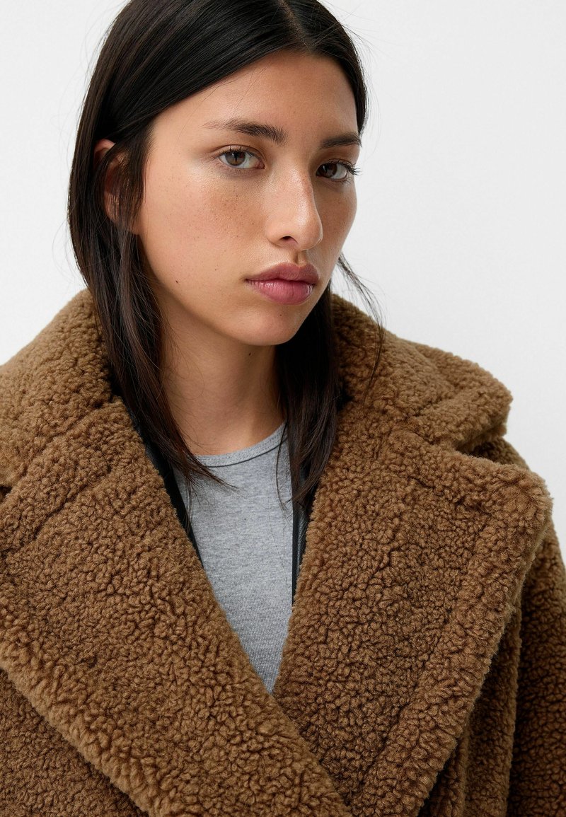 Un manteau brun effiloché avec un large col, fabriqué en matériau doux et duveteux. Porté sur une chemise grise, mettant en valeur sa texture et sa couleur chaude.