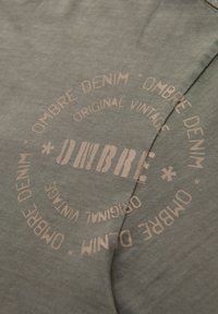 Zelená látka s měkkou strukturou, na které je vytištěn kruhový, vybledlý růžový motiv s nápisem "OMBRE DENIM ORIGINAL VINTAGE" a hvězdami.