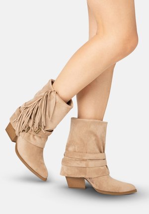Beige ruskind ankelstøvler med stabelhæle, blød skaft, frynsedetalje på den ene støvle, båret på krydsede ben mod ensfarvet baggrund.