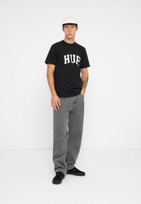 Fekete pamut póló fehér "HUF" logóval, laza szürke farmernadrággal és fekete sportcipővel párosítva. A modellen fehér sapka van, háttérben fehér fal áll.