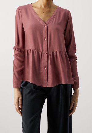 Femme portant un chemisier rose poudré à manches longues avec boutons, une taille froncée et un pantalon noir, debout devant un fond clair uni.