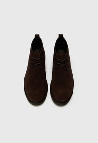 Botas de tobillo de ante marrón con punteras redondas, cordones planos y una superficie texturizada. Diseño minimalista con suelas oscuras a juego.