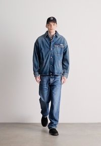 Τζάκετ από τζιν με κουμπιά μπροστά, τσέπη στο στήθος και μπλε τζιν. Το μοντέλο φοράει μαύρα αθλητικά παπούτσια και μαύρο καπέλο. Ο φόντος είναι λευκός.