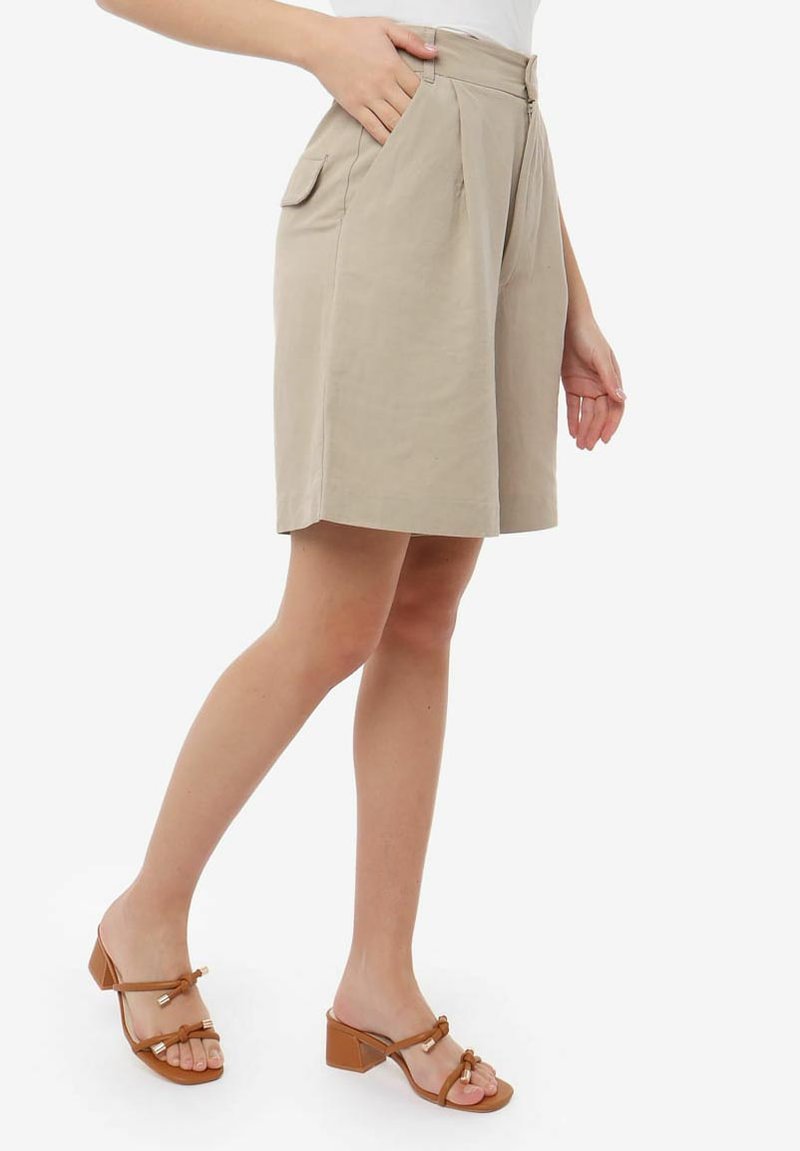Pantaloni corti beige su misura con pieghe, tasche laterali e vita con passanti per la cintura. Abbinati a sandali con tacco quadro aperti davanti color marrone.